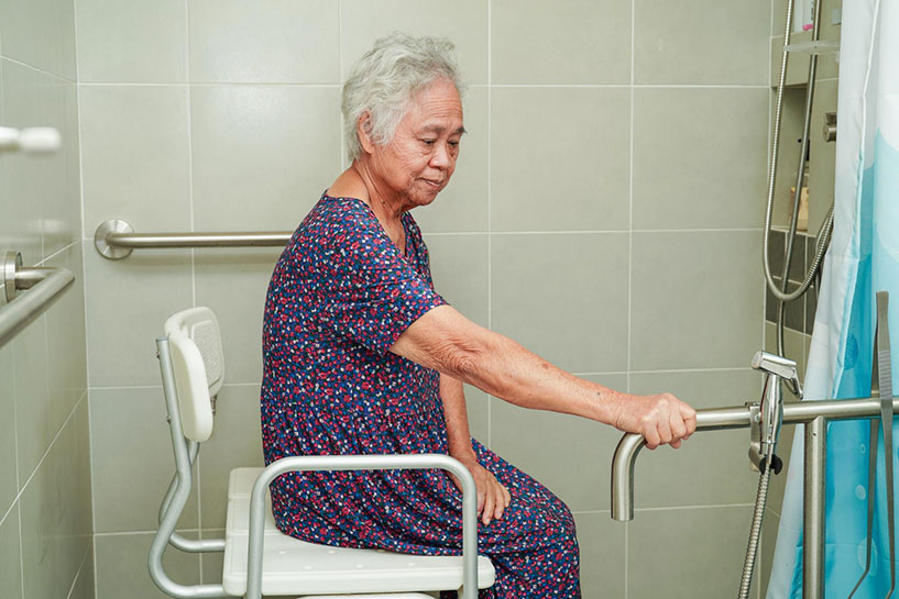 Senior woman sitting on chair & holding handrail safety grab bar in ADA bathroom